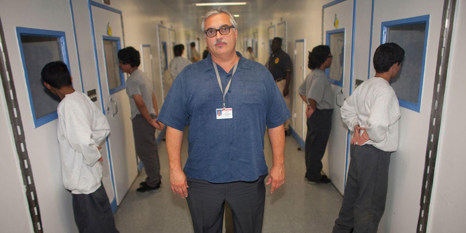 Man stands in the middle with name tag, children behind him stand facing row of doors, hands on their backs. 