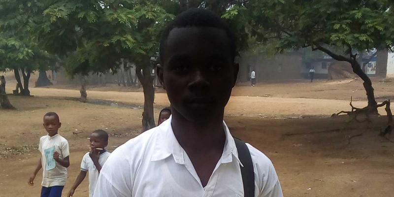Black boy in school uniform standing on yard, two children pass by behind him.