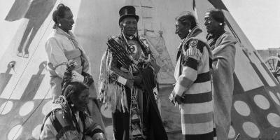 A group of older men dressed in First Nations regalia stand together outside a tipi.